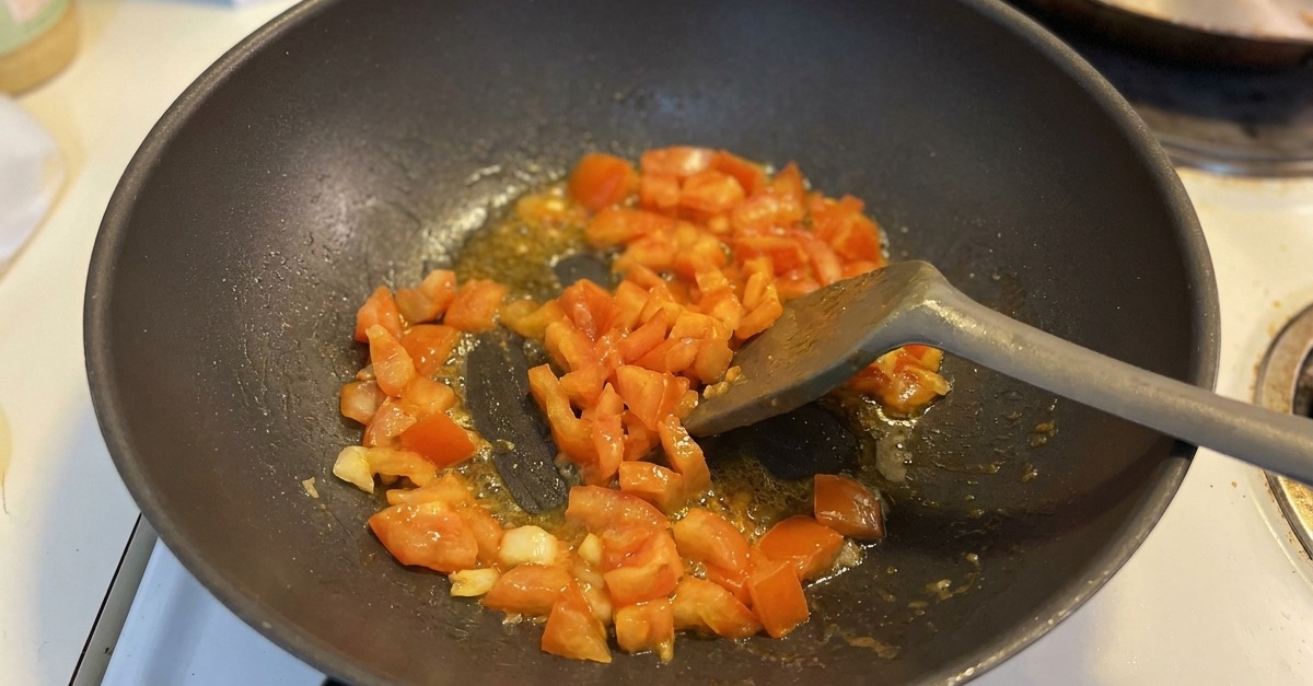 Dés de tomates revenant avec de lail dans un wok sombre, le jus commençant à bouillonner au fond.