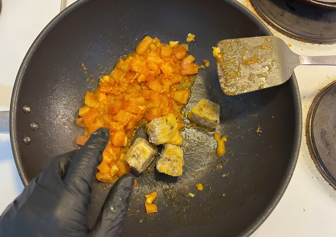 Cubes surgelés de pulpe de fruit de la passion et piments hachés ajoutés à une poêle de tomates par une main gantée.