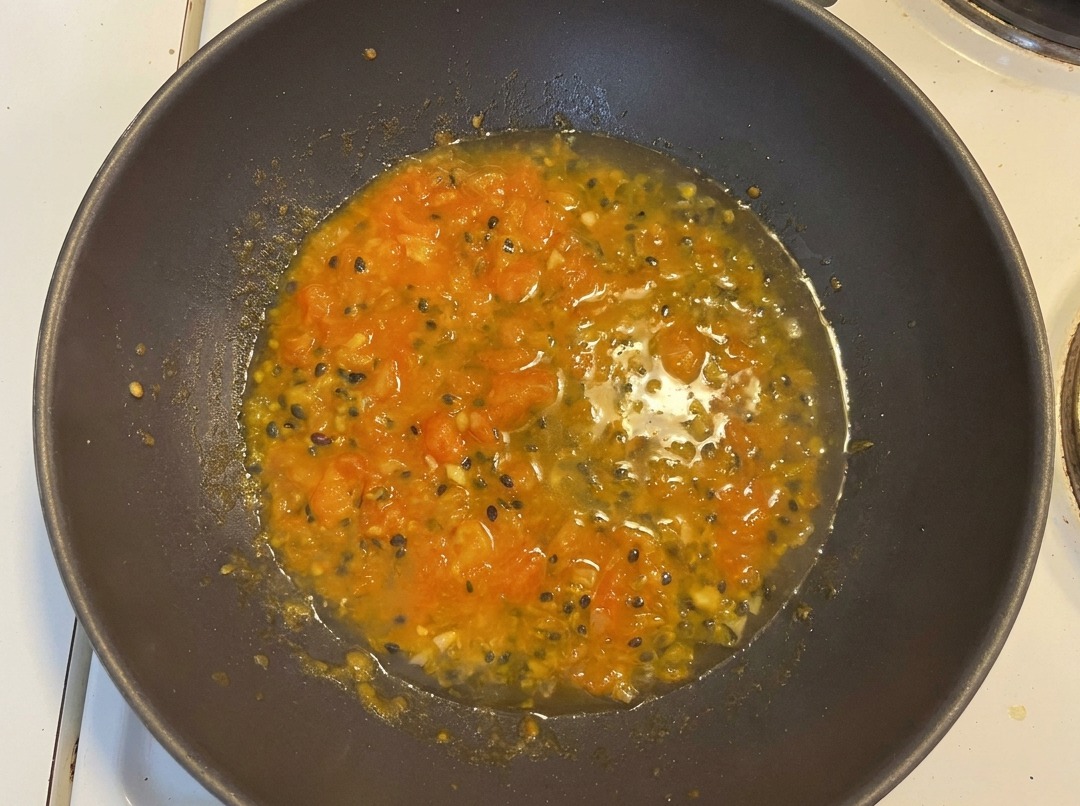 Eau ajoutée au mélange de tomates et fruits de la passion dans un wok, créant une base de soupe orangée avec des graines visibles.