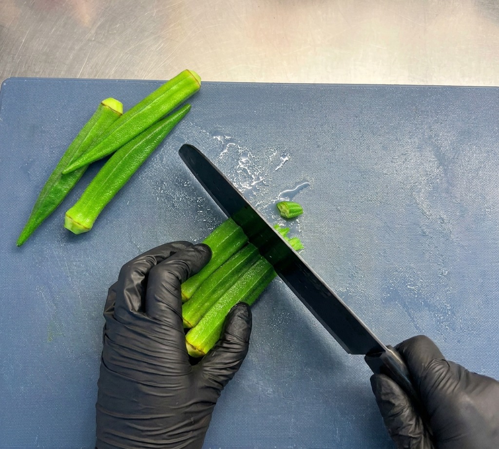 Personne avec des gants noirs utilisant un couteau de chef pour couper le gombo vert en petites rondelles sur une planche bleue.