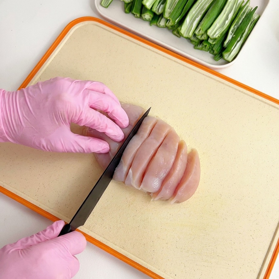 Blanc de poulet cru tranché avec un couteau noir sur une planche à découper claire par des mains gantées.