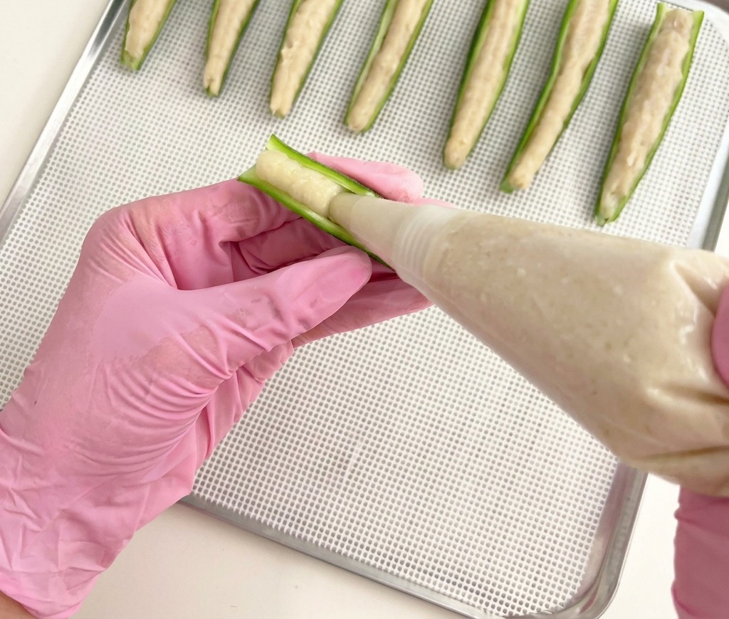 Une personne utilisant une poche à douille pour garnir des moitiés dokra vert avec une pâte de poulet sur une grille métallique.