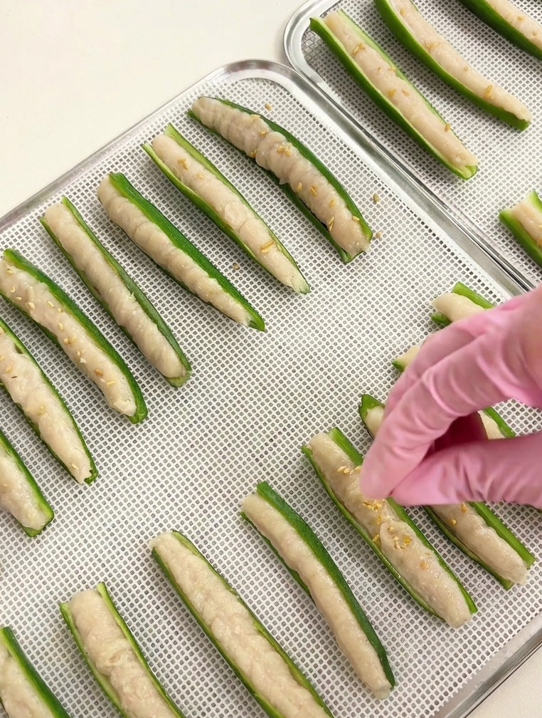 Gros plan dune main saupoudrant des graines de sésame blanc sur des okras farcis sur un plateau argenté.