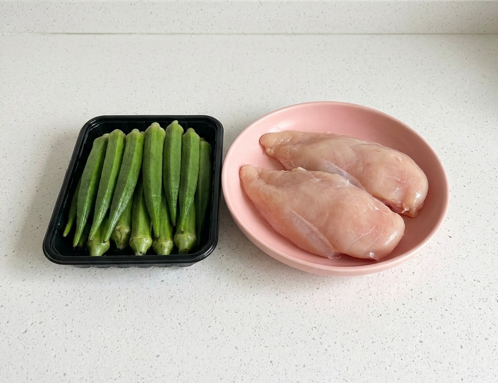 Un plateau noir de gombo frais posé à côté dune assiette contenant deux blancs de poulet crus sur un plan de travail blanc.