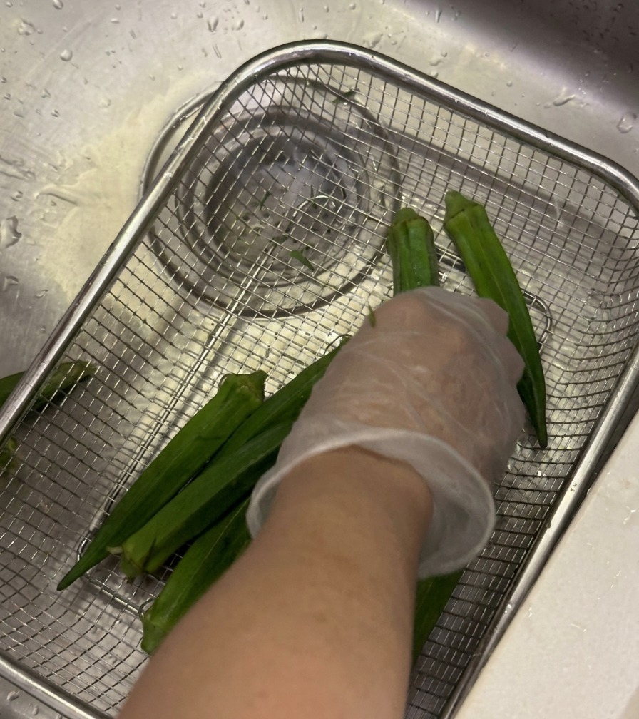Gousses de gombo frais lavées dans un panier sous le robinet avec une main gantée frottant les gousses.