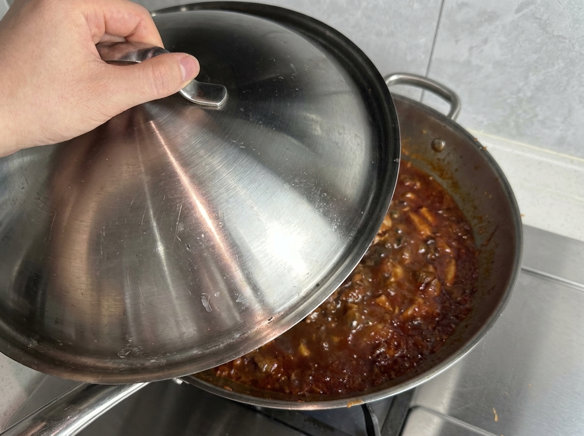 Une main posant un couvercle en inox sur une grande poêle remplie de sauce bolognaise bouillonnante.