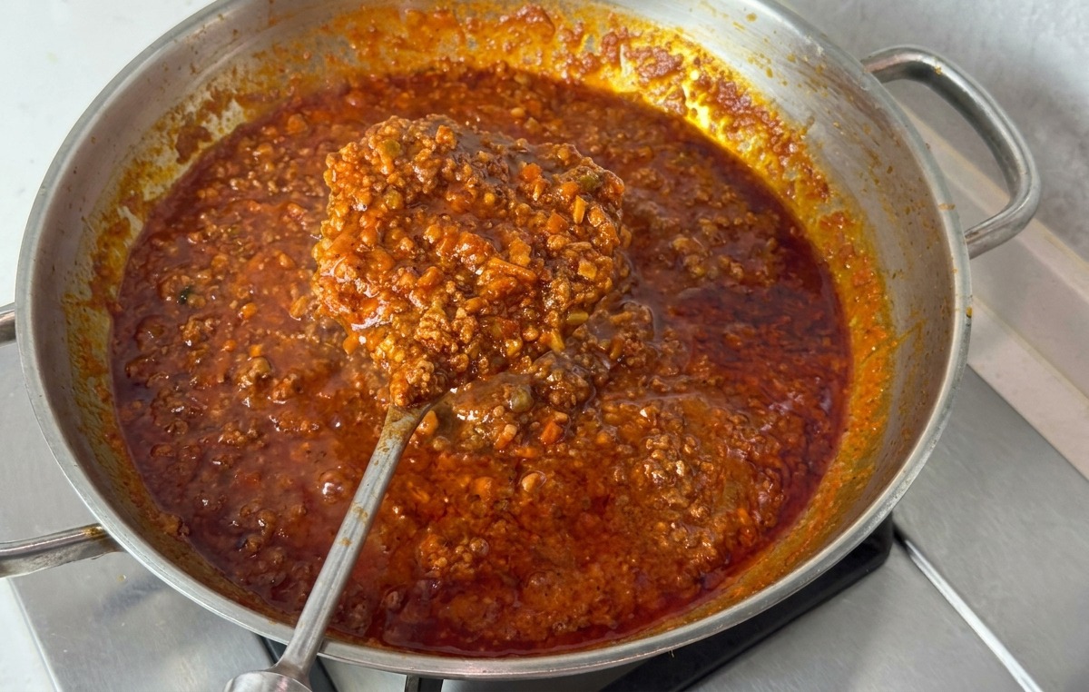 Poêle en inox remplie dune sauce bolognaise épaisse rouge foncé soulevée par une grande cuillère en métal.