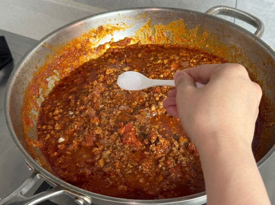 Une main tenant une petite cuillère en plastique blanc remplie de sel fin au-dessus de la sauce bouillonnante.