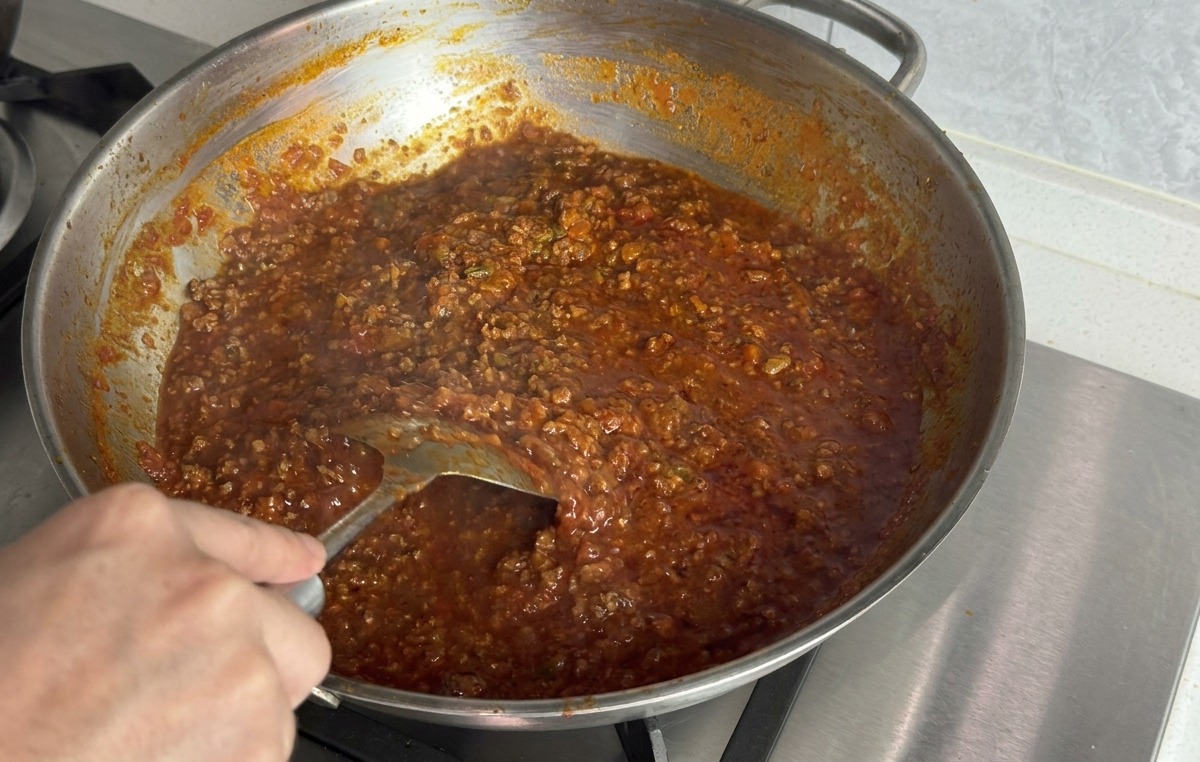 Une main utilisant une spatule en métal pour incorporer le beurre fondant dans la sauce bolognaise épaisse.