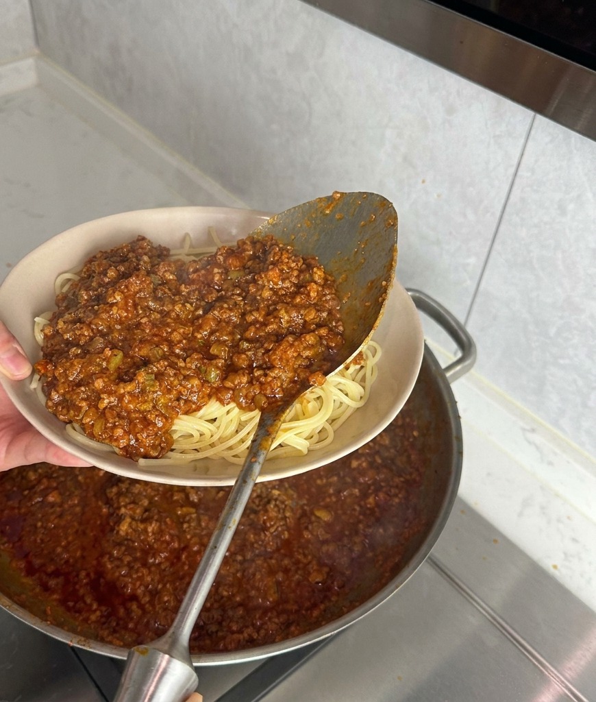 Une louche en métal versant une sauce bolognaise épaisse sur un lit de spaghettis nature dans un bol beige.
