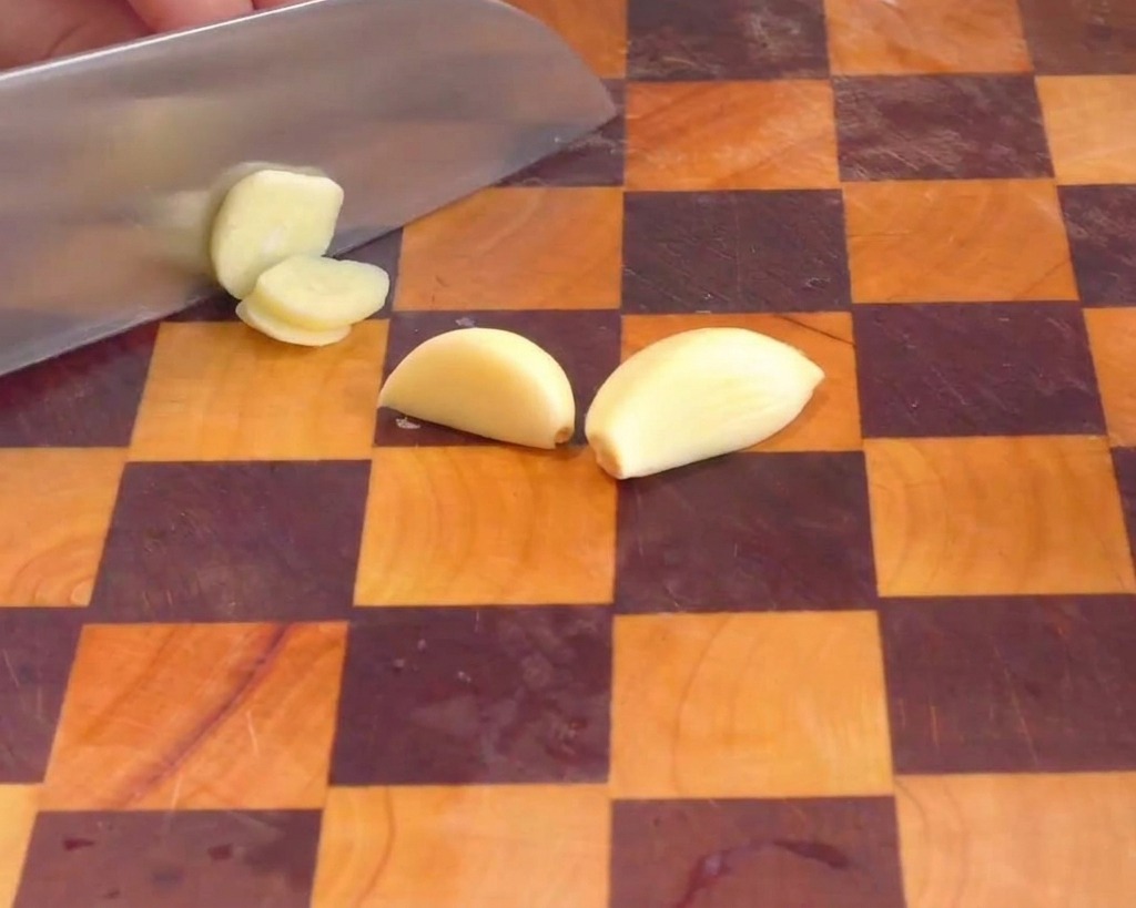 Une main utilisant un couteau de chef pour couper des gousses dail pelées en fines lamelles.