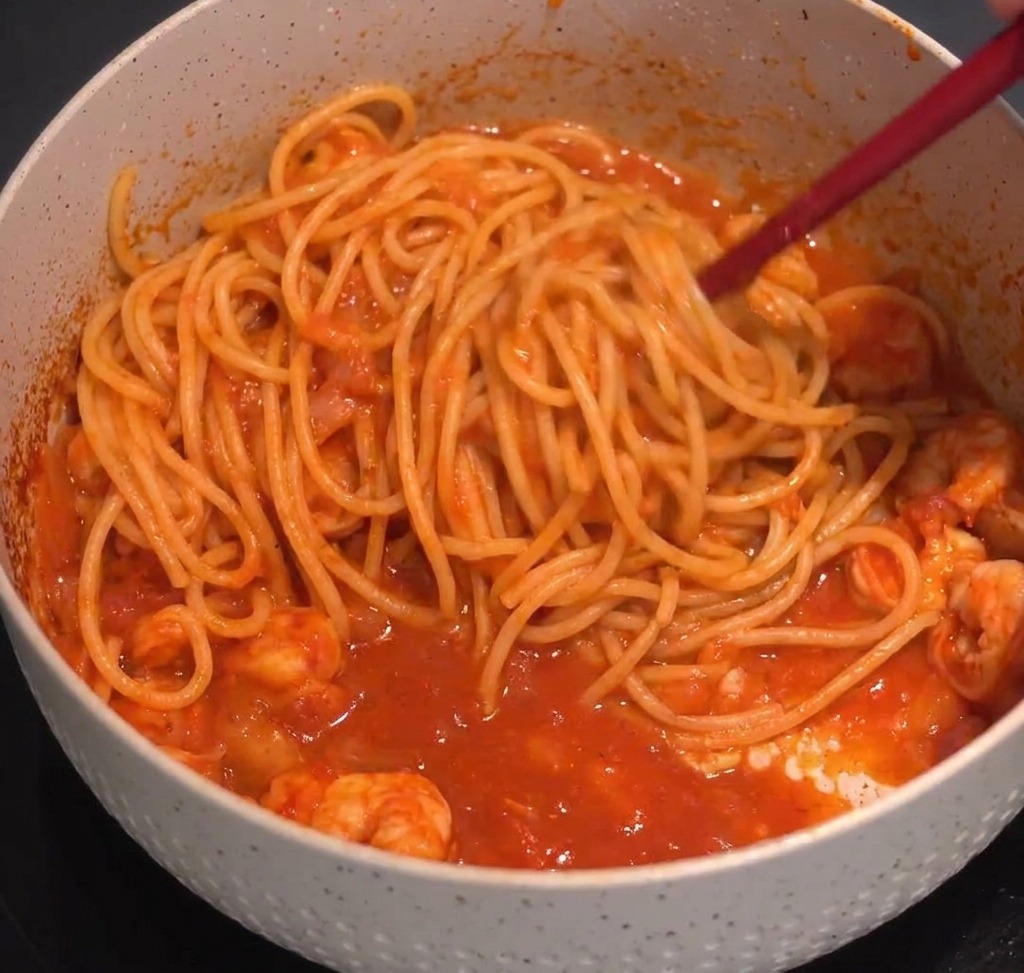 Spaghetti mélangés à une sauce rouge aux crevettes à laide dune pince de cuisine.