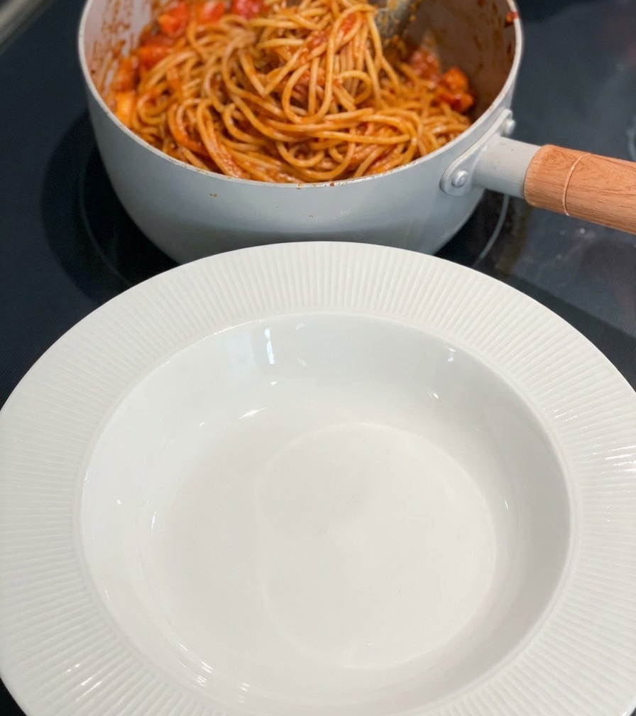 Une poêle de pâtes aux crevettes à côté dun bol blanc vide sur la plaque de cuisson.