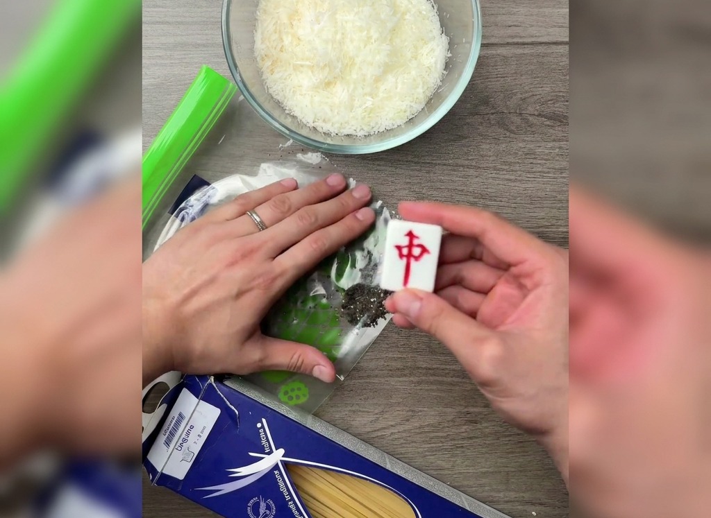 Une main utilisant le côté plat dune pièce de mah-jong pour écraser des grains de poivre noir dans un sac en plastique sur une table en bois.