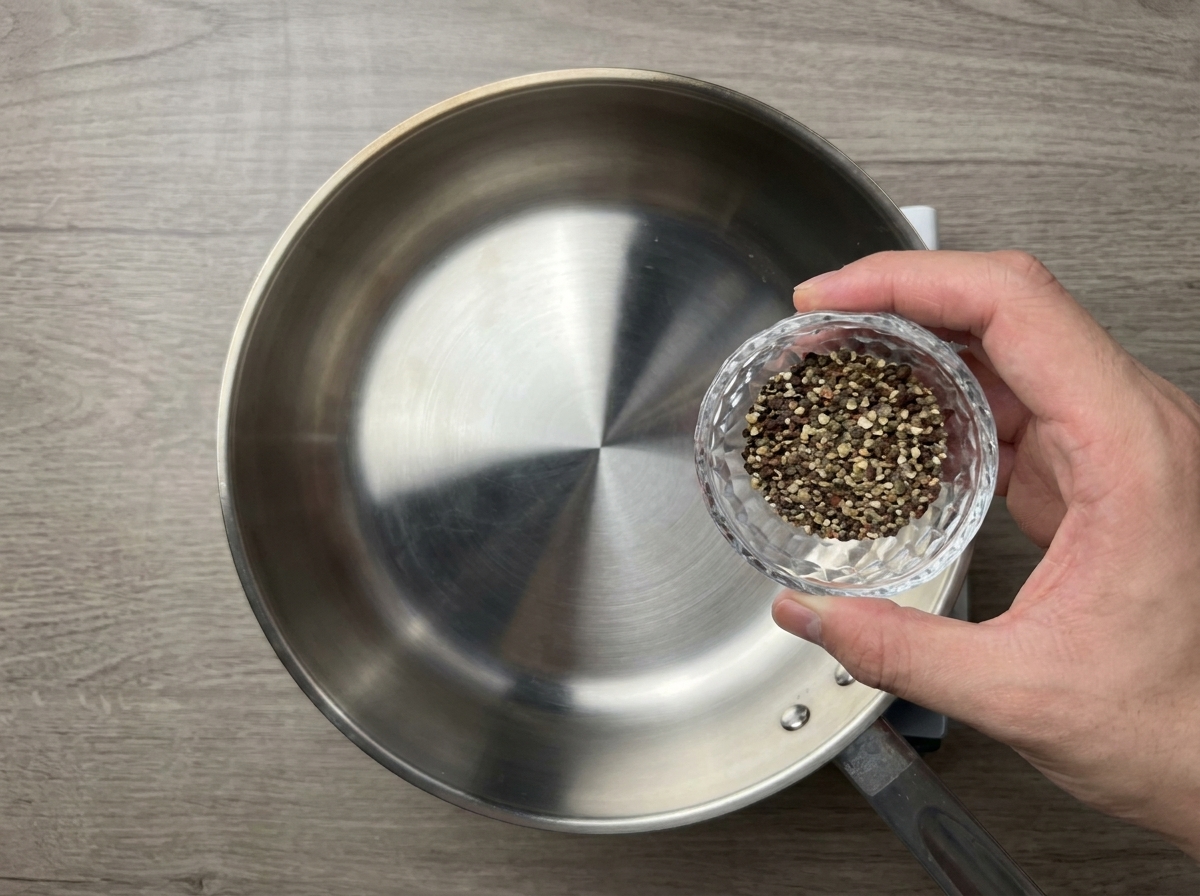 Poivre noir concassé grossièrement versé dun petit bol en verre dans une poêle en inox préchauffée à sec.