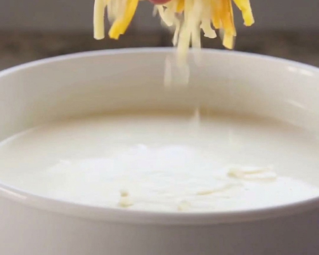 Une main déposant du fromage râpé orange et blanc dans une sauce crémeuse chaude