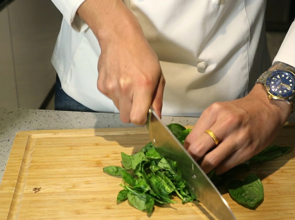 Mains utilisant un grand couteau pour hacher des feuilles de basilic frais sur une planche en bois.