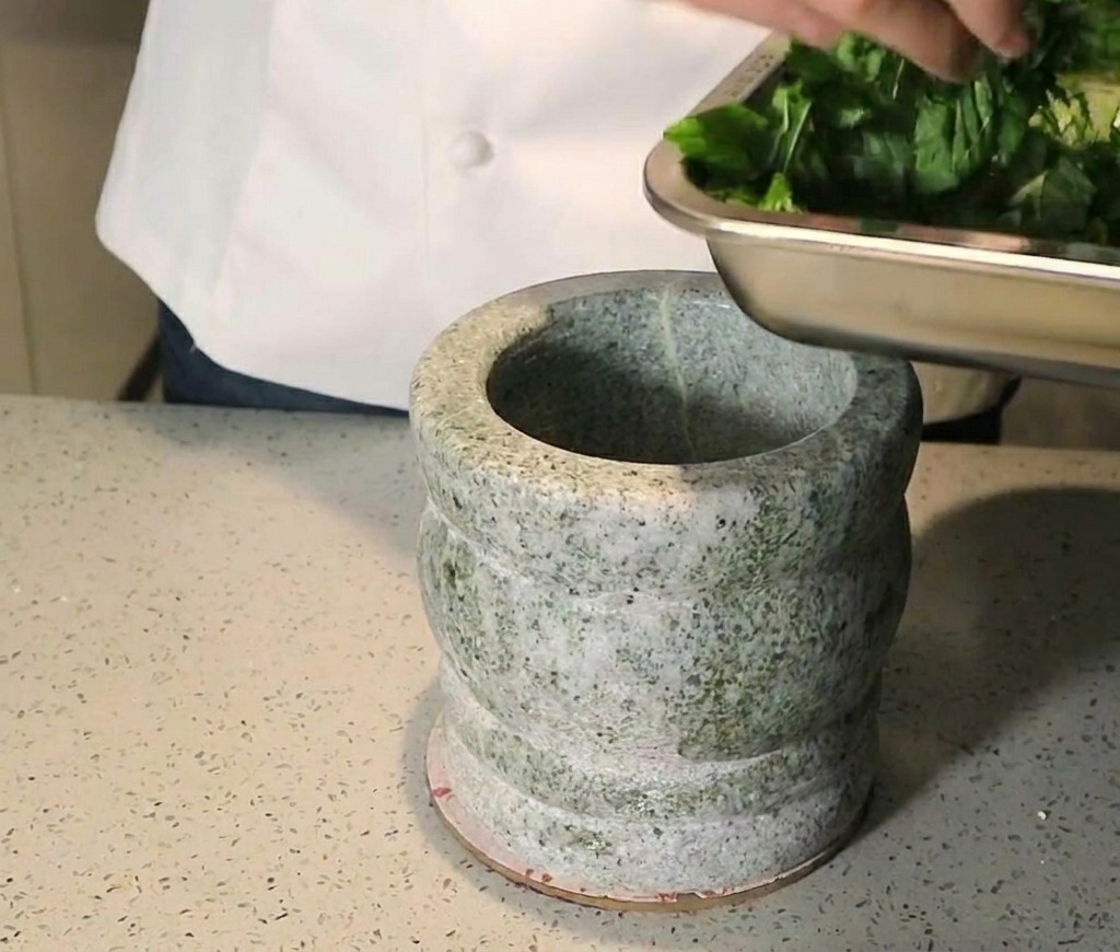 Une personne ajoutant des feuilles de basilic hachées dans un mortier en granit lourd.