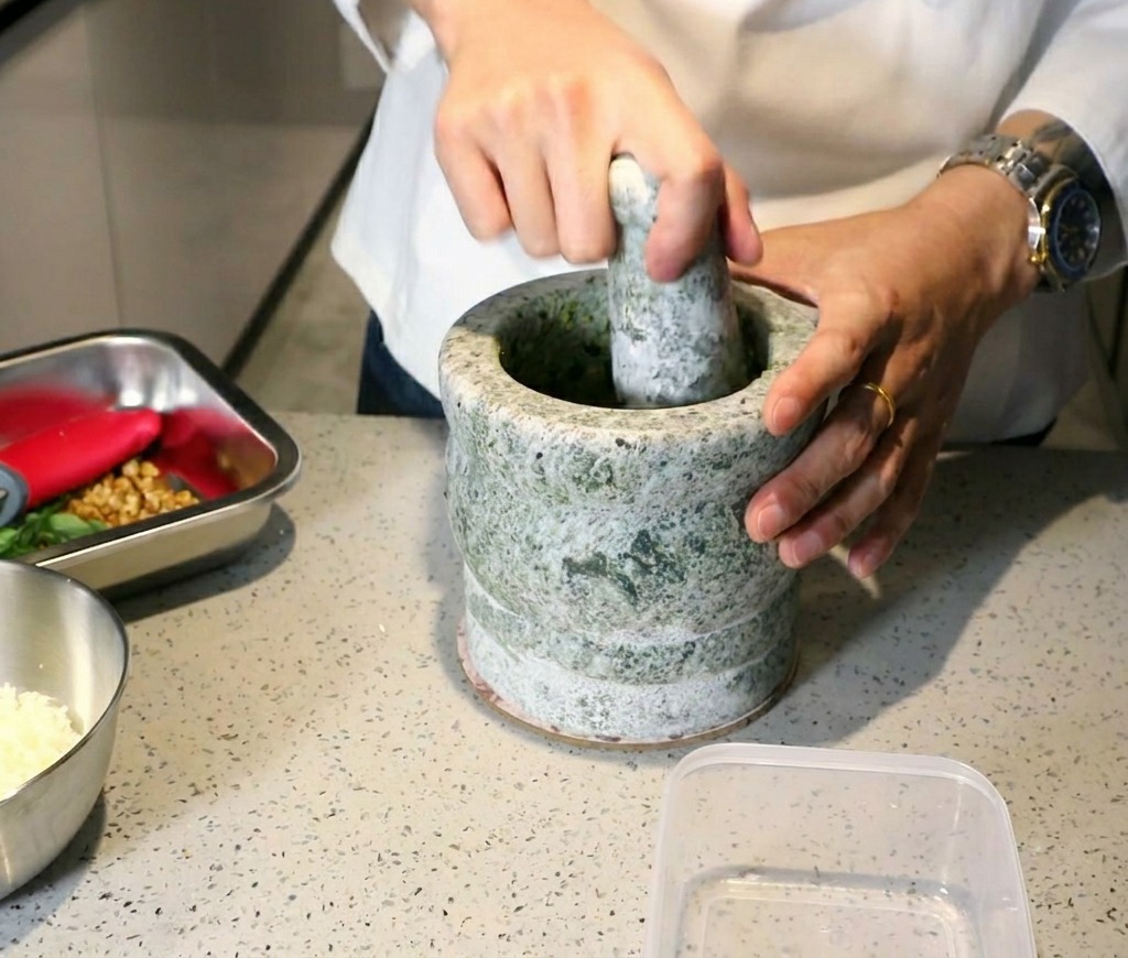 Un chef utilisant un mortier et un pilon en pierre pour transformer le basilic et lail en une pâte de pesto épaisse.