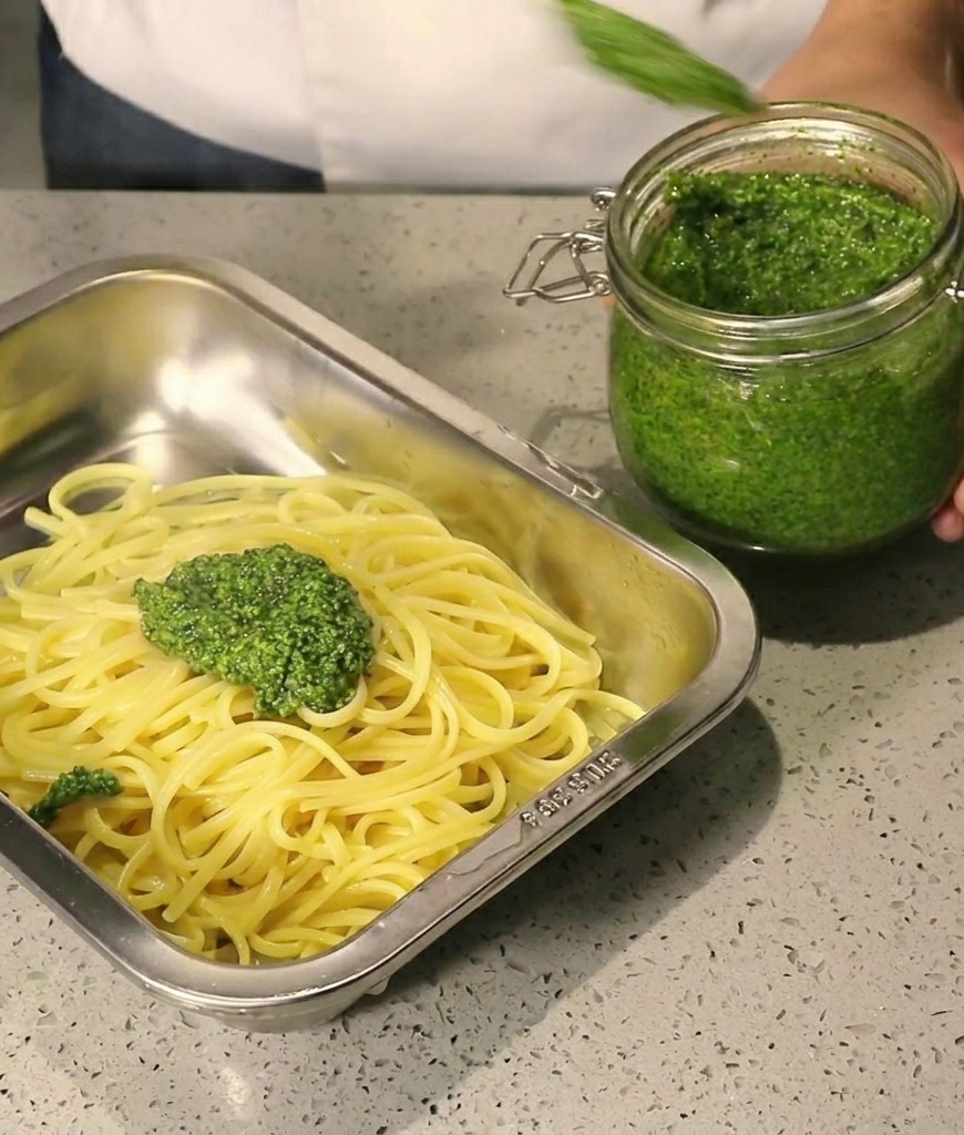 Grosses cuillères de pesto vert déposées sur des spaghettis chauds dans un plat en inox.