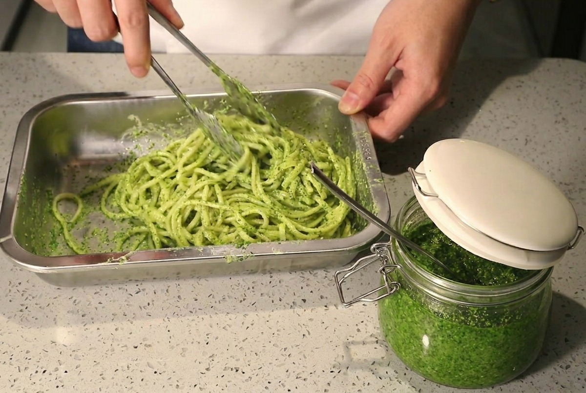 Utilisation de pinces pour mélanger les spaghettis avec le pesto et la crème dans un plat en métal.