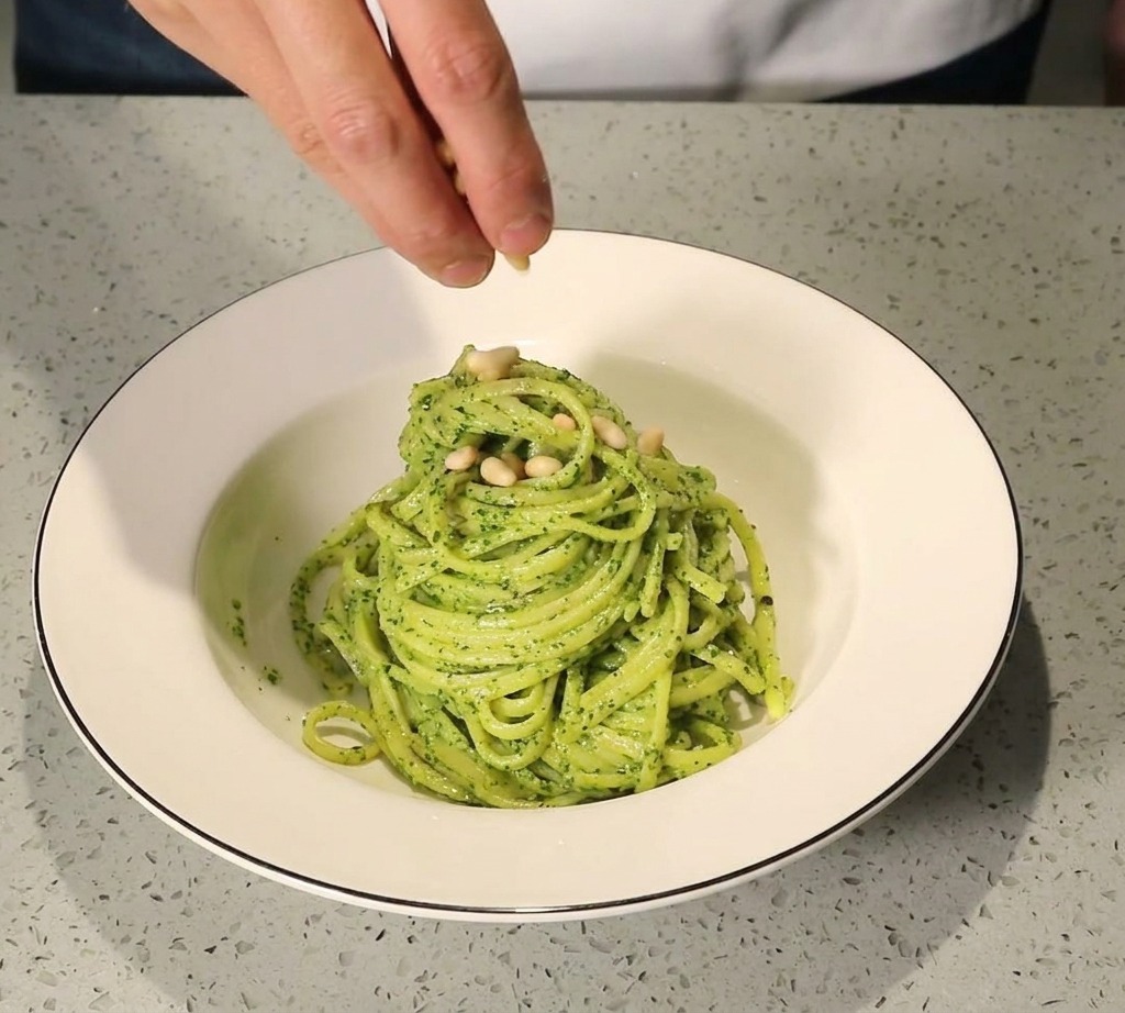 Une main parsemant des pignons de pin dorés sur une portion de pâtes au pesto dans un bol blanc.