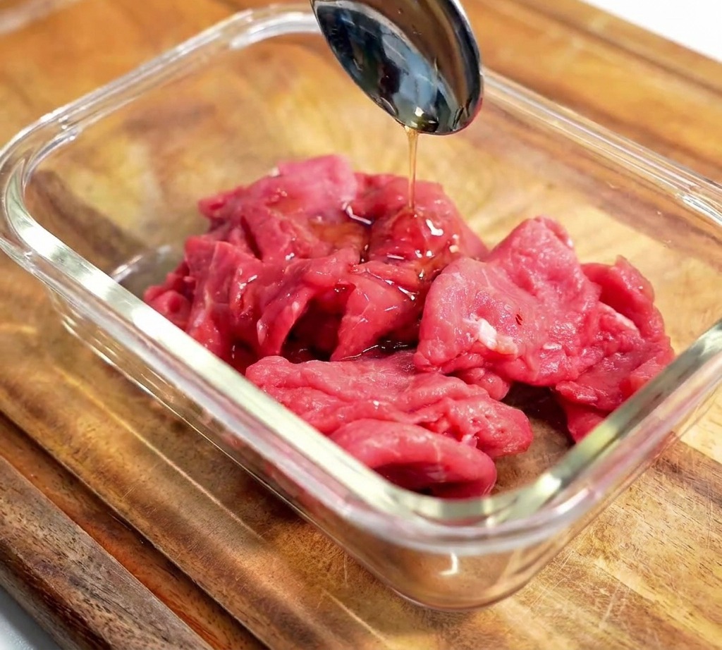 Une cuillère versant un assaisonnement sombre sur des tranches de bœuf cru dans un bol en verre.