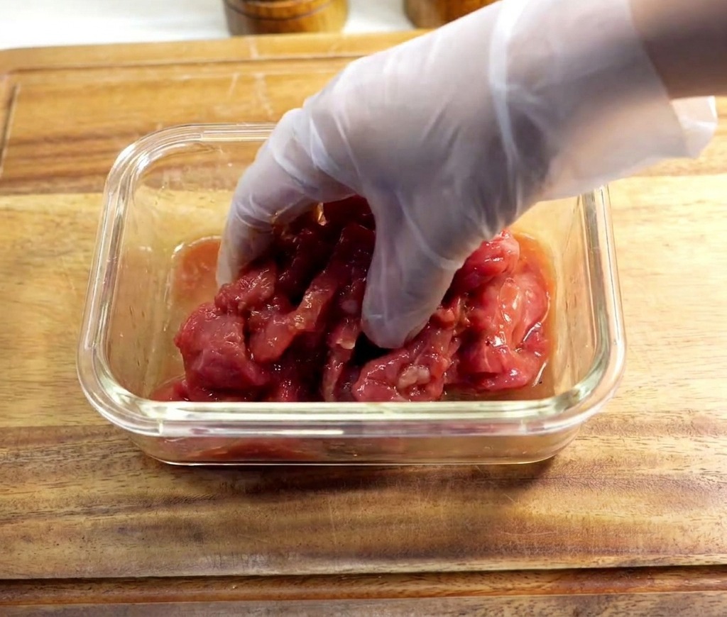 Une personne portant un gant mélangeant des tranches de bœuf mariné dans un bol en verre.