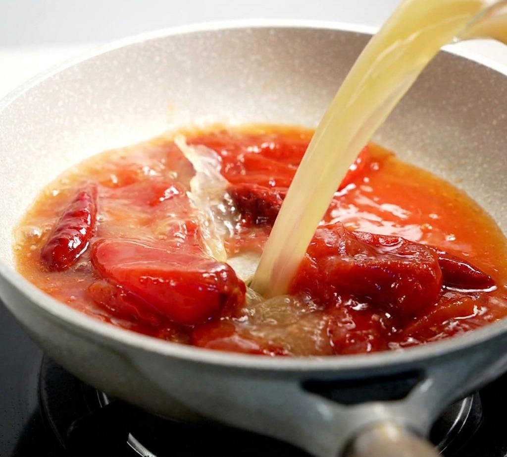 Un bouillon clair versé dans une poêle contenant un mélange de sauce tomate bouillonnant.