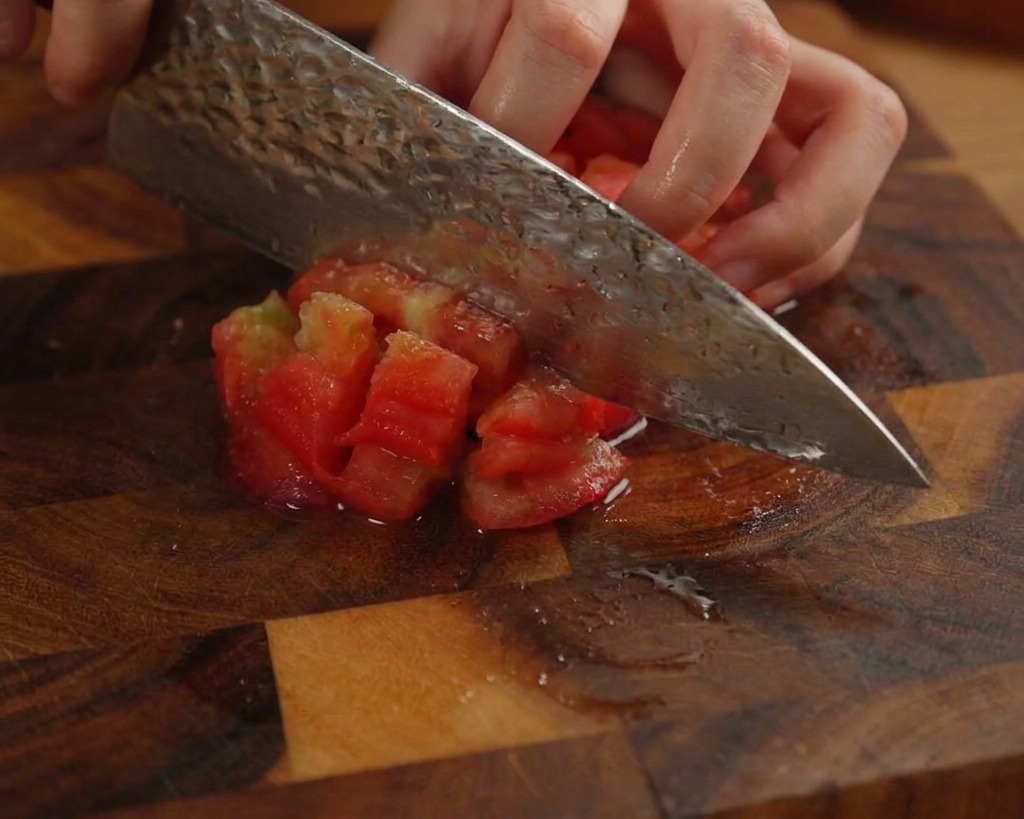 Une personne coupant une tomate pelée en petits cubes sur une planche en bois sombre.