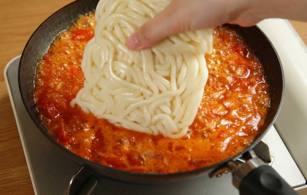 Une main déposant un bloc blanc de nouilles udon dans une poêle de sauce tomate frémissante.