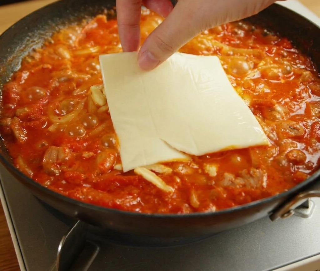 Une main posant une tranche carrée de fromage jaune sur la mozzarella fondante et les nouilles.