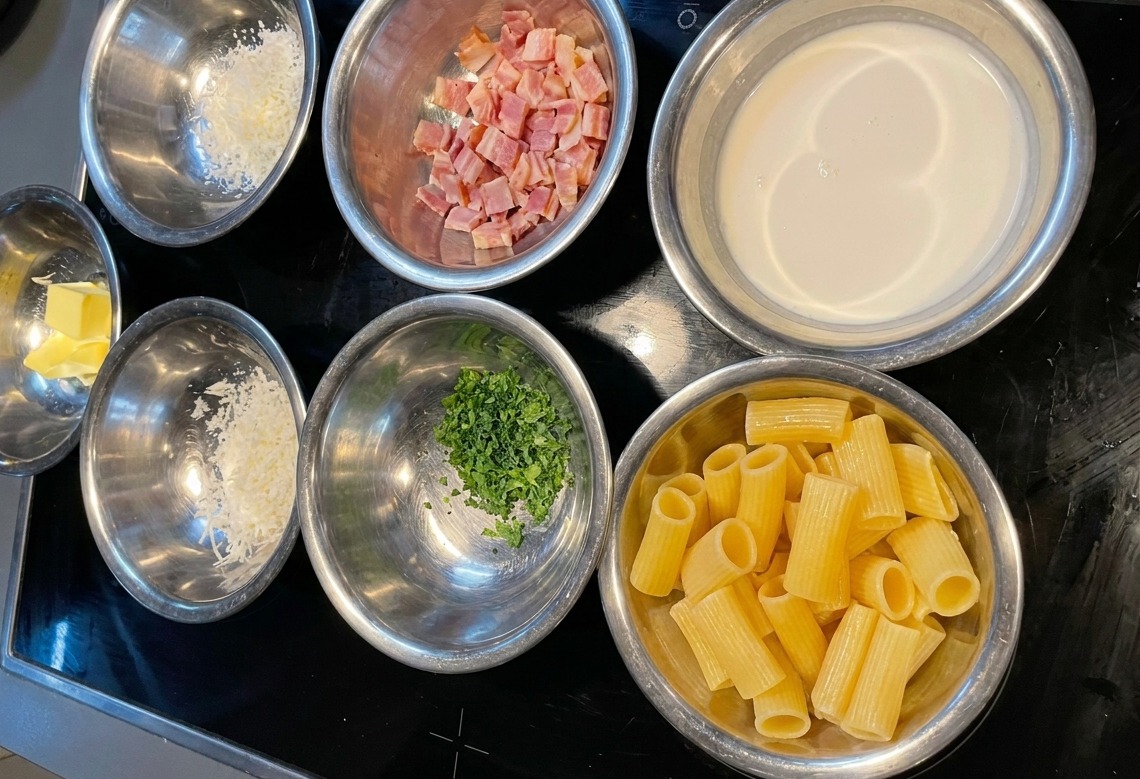 Plusieurs bols en inox contenant les rigatoni, la crème, les lardons, le fromage et le persil disposés sur un plan de travail sombre.