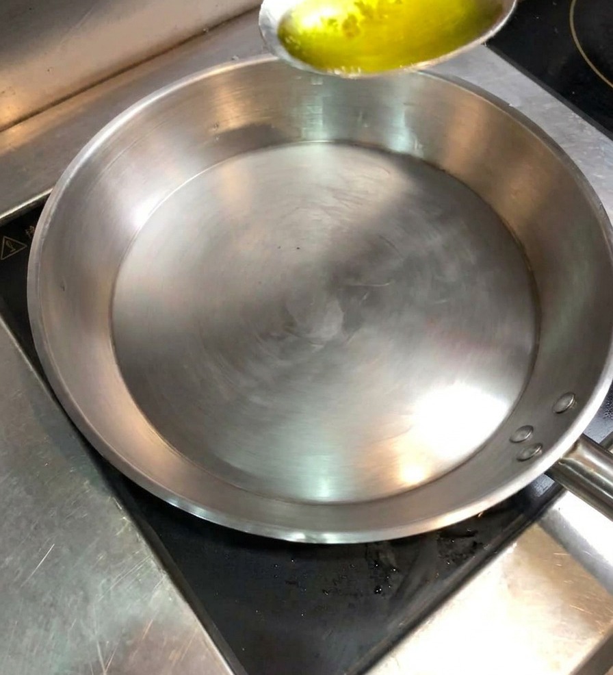 Une cuillère versant de lhuile de cuisson dorée dans une poêle en inox bien chaude.