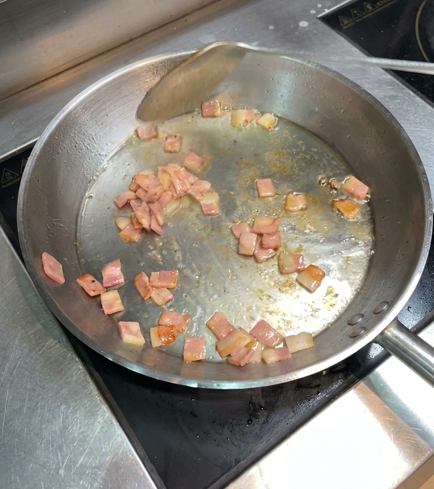 Morceaux de bacon qui grésillent et dorent dans la poêle, remués avec une spatule métallique.