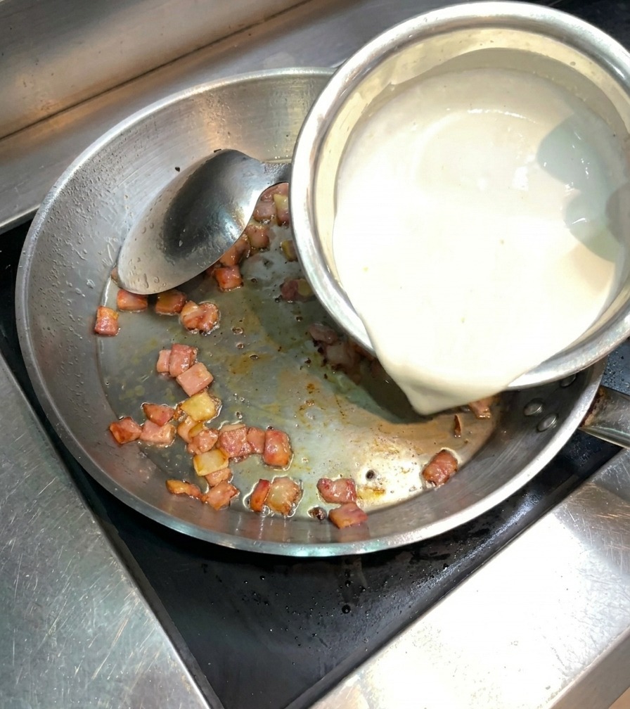 Crème liquide épaisse versée dans une poêle en inox contenant le bacon croustillant.