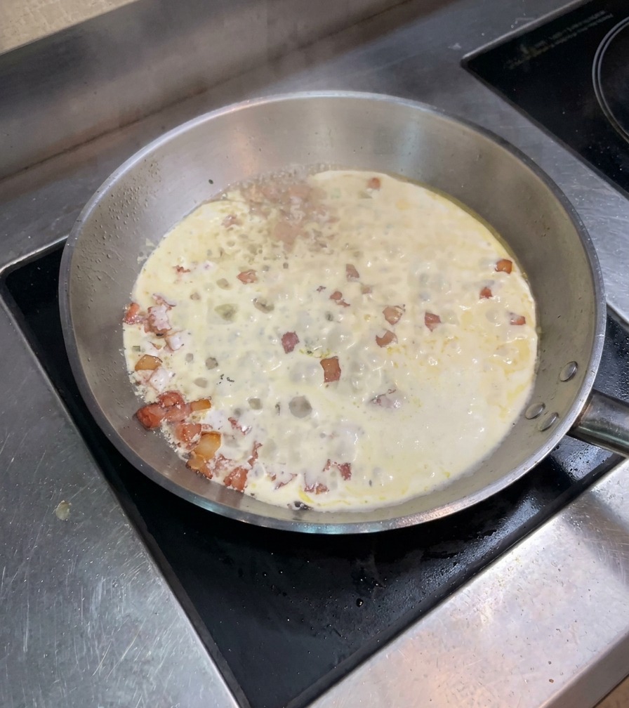 Mélange de crème et de lardons frémissant et bouillonnant dans une large poêle sur la cuisinière.