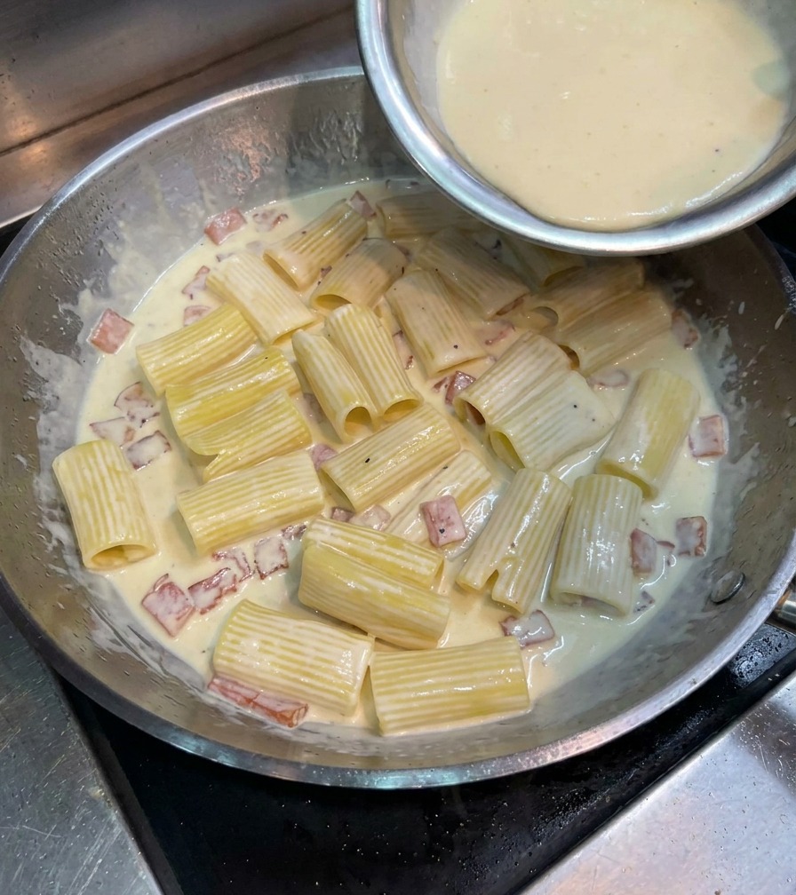 Mélange jaune dœuf et fromage versé sur les rigatoni dans une grande poêle métallique.