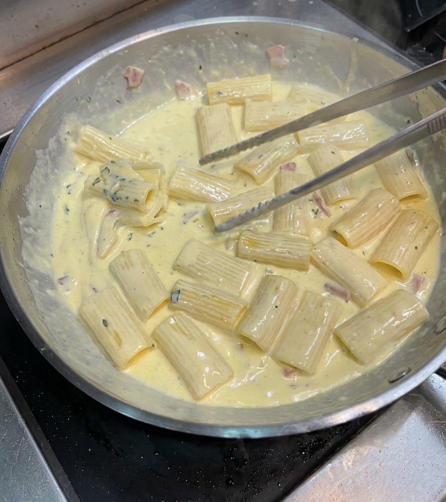 Pince mélangeant les rigatoni dans la poêle pour bien les enrober de la sauce carbonara épaisse.