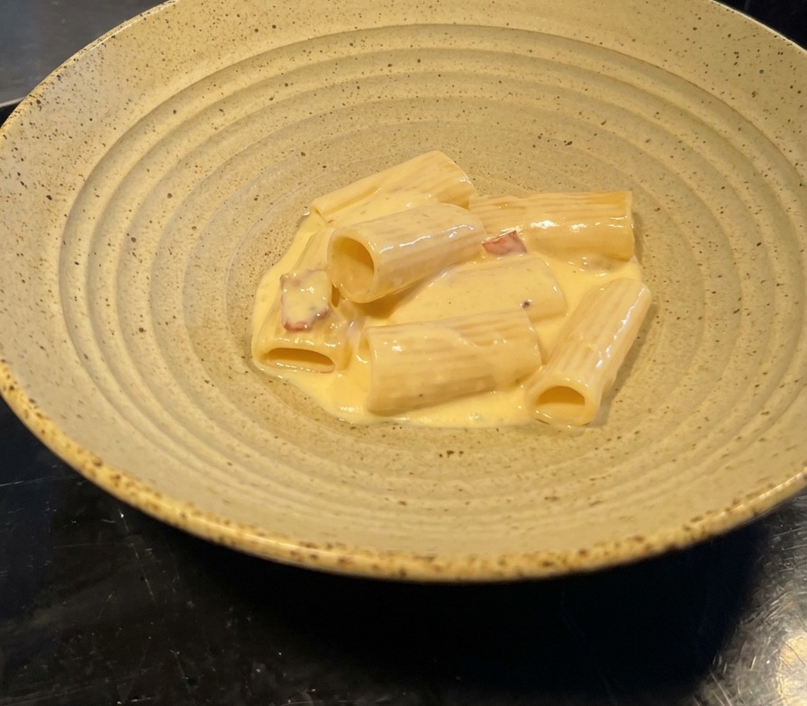Portion de rigatoni nappée de sauce jaune pâle avec des éclats de bacon, servie dans un bol en céramique texturé.