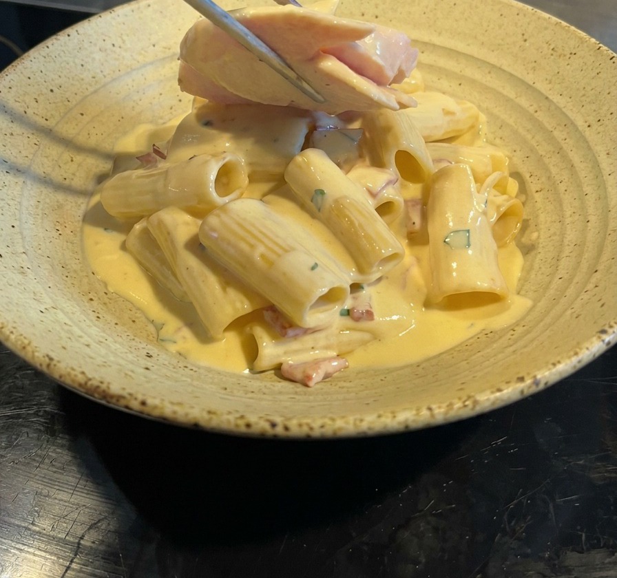 Pince déposant des tranches de blanc de poulet grillé sur une portion de rigatoni carbonara au bacon.
