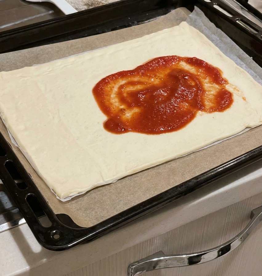 De la sauce tomate rouge épaisse déposée au centre dune pâte à pizza rectangulaire crue sur une plaque.