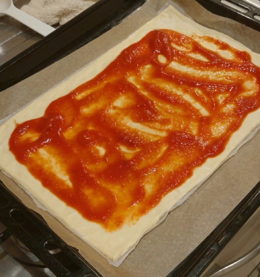 Une base de pizza rectangulaire avec de la sauce tomate étalée uniformément, laissant un bord de pâte libre.