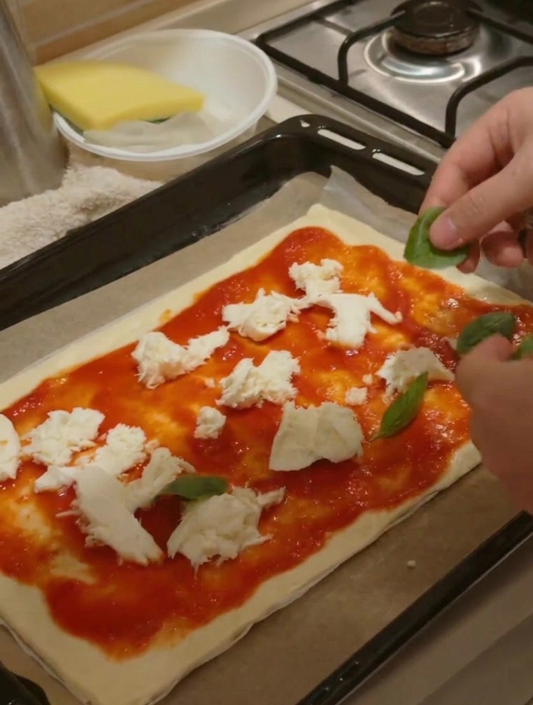 Des mains déposant des feuilles de basilic vert sur une pizza déjà garnie de sauce et de morceaux de fromage.
