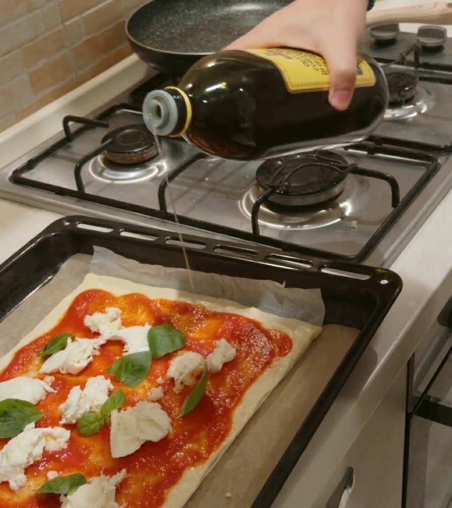 Une main versant un filet dhuile dolive dune bouteille en verre sur une pizza crue garnie de sauce tomate, mozzarella et basilic.