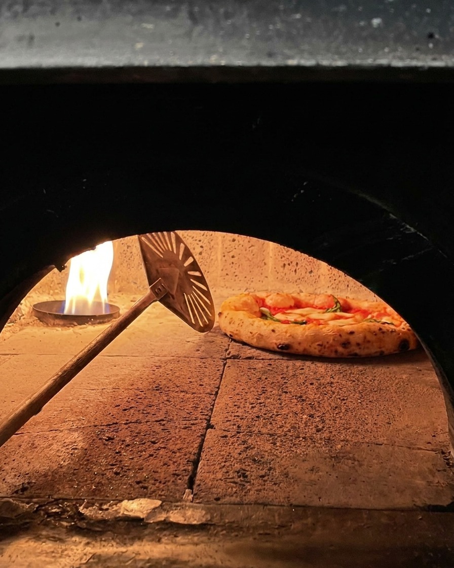 Pizza Margherita cuisant dans un four à bois, tournée par une petite pelle métallique près des flammes.