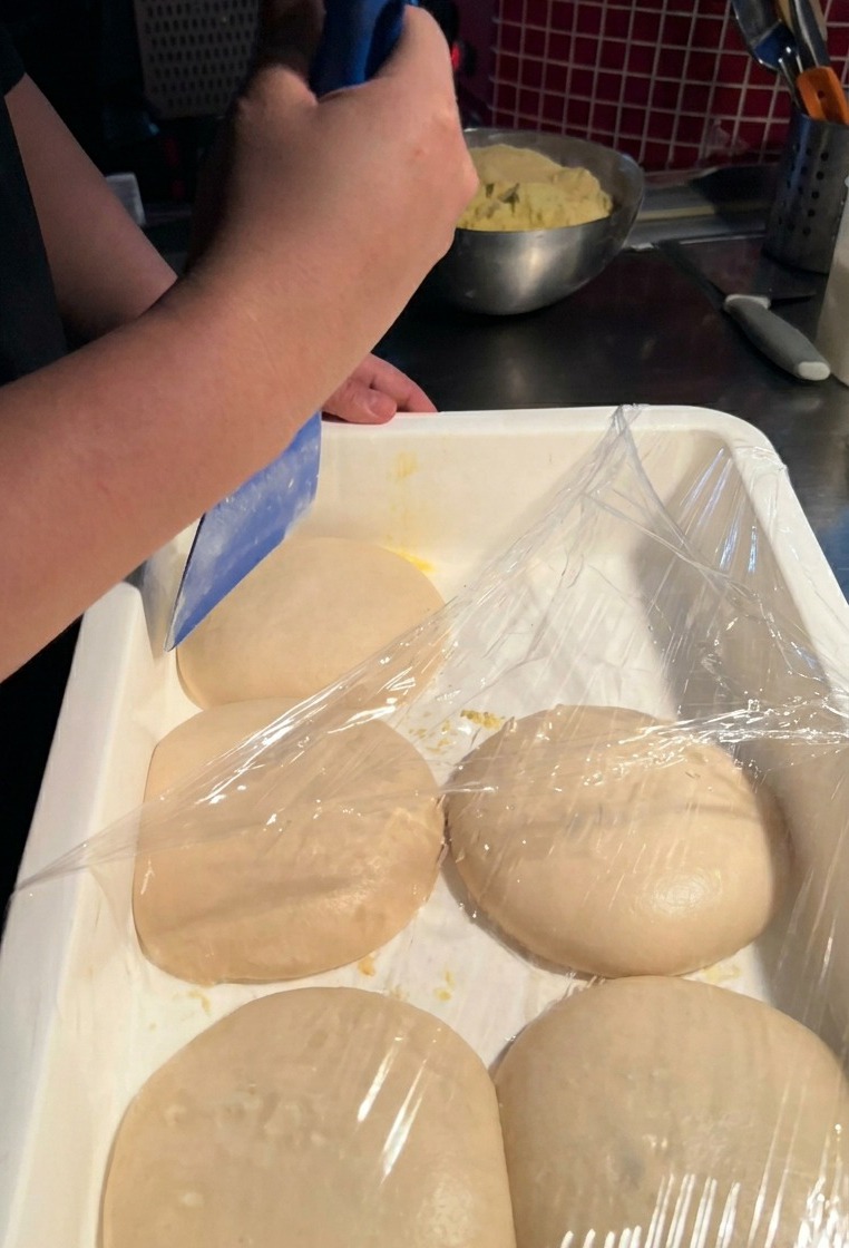 Une personne utilisant un coupe-pâte en plastique bleu pour soulever délicatement une boule de pâte lisse dun bac blanc.