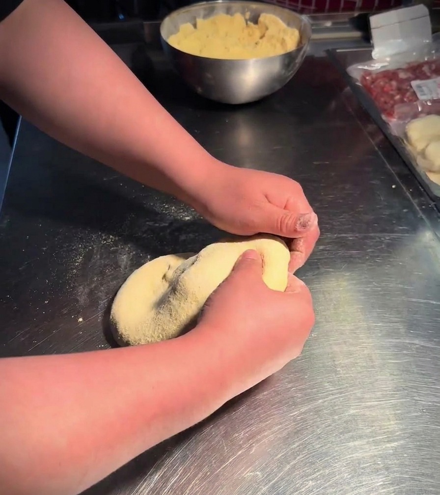 Deux mains pressant et retournant une boule de pâte souple dans un bol en inox rempli de farine de maïs jaune.