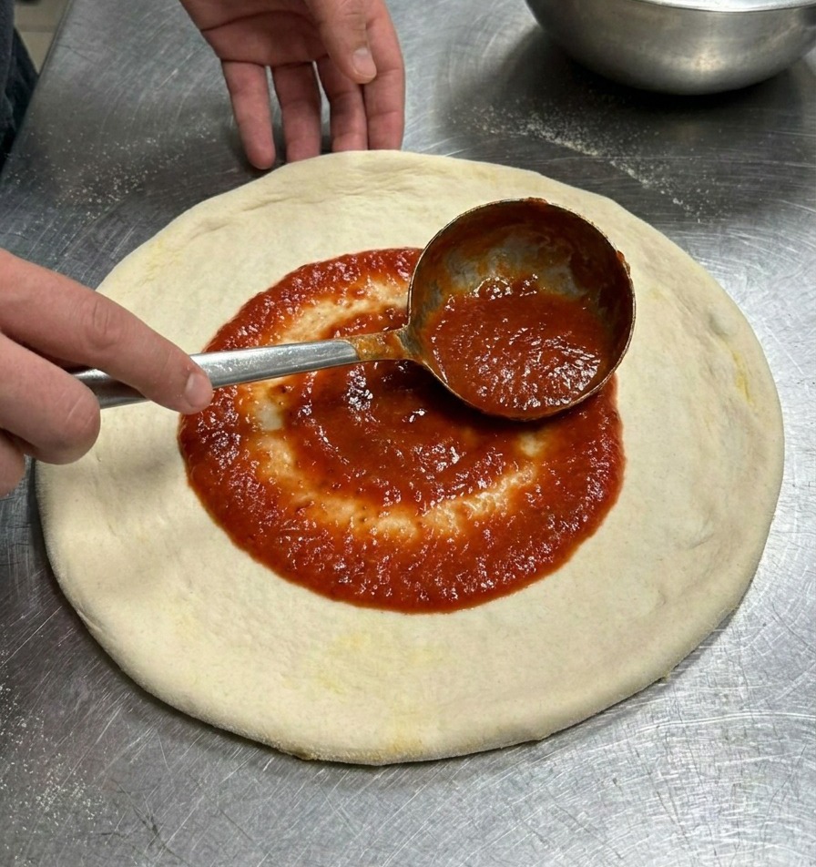 Une louche en métal étalant une sauce tomate rouge vif en mouvement circulaire sur un disque de pâte à pizza.