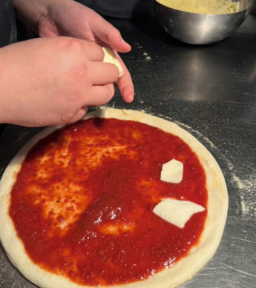 Une personne déposant des morceaux de mozzarella blanche déchirés à la main sur une base de pizza déjà garnie de sauce rouge.
